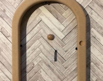 Puede incluir: Un marco de madera arqueado de color beige con un pequeño pomo de madera redondo y un pestillo de metal. El marco está montado sobre un fondo con diseño de espiga marrón claro y gris. El marco tiene un pequeño agujero en el lado derecho.