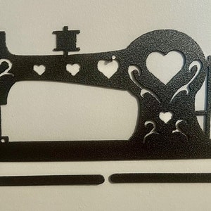 May include: Black metal sewing machine wall decor with a cutout design. The machine has a heart shape in the center and a decorative scroll design. The bottom of the machine has a flat bar for hanging items.