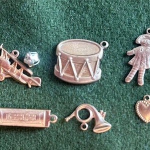 May include: A collection of silver charm ornaments, including a gingerbread man, a toy airplane, a drum, a heart, a musical instrument, and a doll. There are also red and green glass beads.