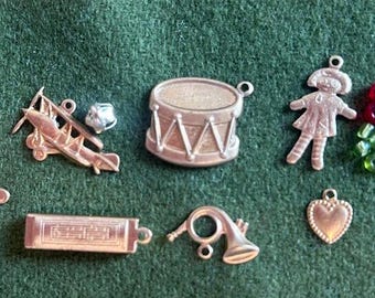 May include: A collection of silver charm ornaments, including a gingerbread man, a toy airplane, a drum, a heart, a musical instrument, and a doll. There are also red and green glass beads.