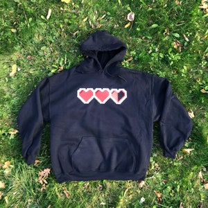 May include: Black hooded sweatshirt with a front pocket and a pixelated heart design. The design features three hearts, with the third heart partially filled. The sweatshirt is made of a soft material and is suitable for casual wear.
