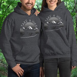Puede incluir: Dos personas con sudaderas con capucha grises con un gr&aacute;fico blanco de monta&ntilde;as y el texto "Take a Hike".