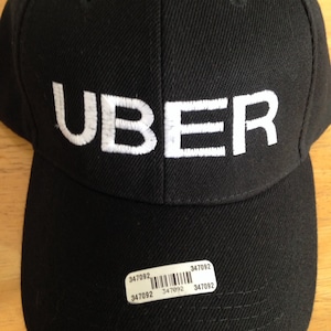 May include: Black baseball cap with the white embroidered word "UBER" on the front. A small sticker with the number "347092" and a barcode is affixed to the brim. The cap is made of a textured fabric.