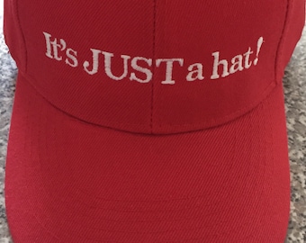 May include: A red baseball cap with white embroidery that reads "It's JUST a hat!"