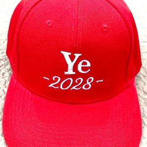 May include: A bright red baseball cap with a curved brim. The cap features the white text "Ye -2028-" embroidered on the front. The cap has a button on top and a structured design.