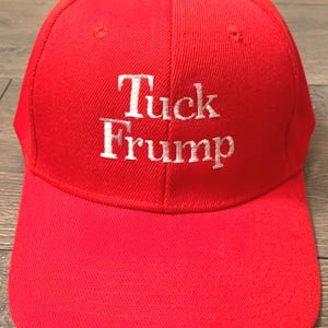 May include: A bright red baseball cap featuring the white embroidered text Tuck Frump centered on the front panel. The cap has a structured crown, a curved brim, and a button at the top, constructed from a durable fabric with visible stitching.