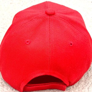 May include: A solid red baseball cap, viewed from the top, showing the crown and the curved bill. The cap has a structured design with a button on top and ventilation eyelets. The fabric has a subtle texture.