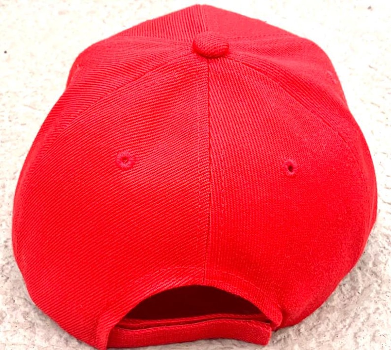 May include: A solid red baseball cap, viewed from the top, showing the crown and the curved bill. The cap has a structured design with a button on top and ventilation eyelets. The fabric has a subtle texture.
