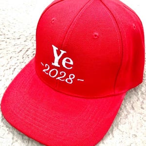 May include: A bright red baseball cap with the word "Ye" and the year "2028" embroidered in white. The cap has a curved brim and a rounded top.