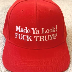 May include: A bright red baseball cap with white embroidered text that reads "Made Ya Look! FUCK TRUMP". The cap has a curved brim and a button on top. The hat is made of a sturdy fabric.