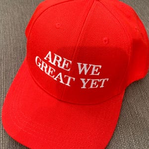 May include: Red baseball cap with white embroidered text that reads "Are We Great Yet".