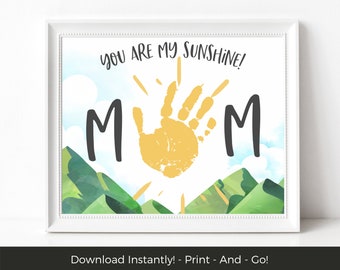May include: A printable art print with a handprint in the center, surrounded by the words "YOU ARE MY SUNSHINE!" and the letters "M" on either side. The handprint is yellow and the background is a watercolor style landscape with green hills and blue sky.
