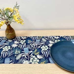 Może przedstawiać: Niebieski bieżnik stołowy z kwiatowym wzorem przedstawiającym białe kwiaty i zielone liście. Na bieżniku stołowym znajduje się niebieski talerz ceramiczny, a w tle stoi mały brązowy wazon z żółtymi kwiatami.