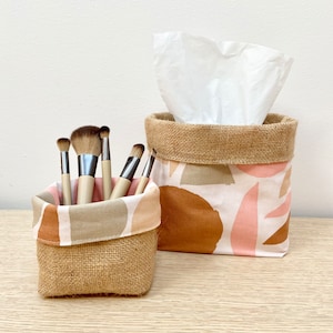 May include: Two fabric storage baskets with burlap trim. One basket is filled with makeup brushes and the other holds a box of tissues. The baskets have a white background with abstract pink, brown, and green shapes.
