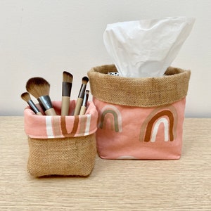 May include: Two fabric storage containers. One holds makeup brushes, the other tissues. Both are made of burlap and a pink fabric with rainbow patterns. The containers sit on a light wood surface.