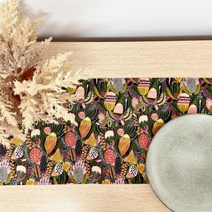 May include: A table runner with a floral pattern featuring a variety of colourful flowers on a dark green background. The runner is placed on a light wood table with a grey ceramic plate.