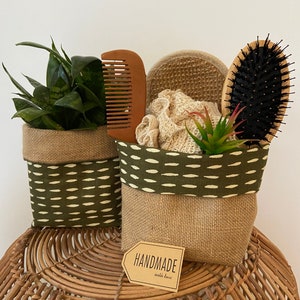 May include: Two decorative baskets made of burlap and olive green fabric with white accents. One basket holds a small plant, the other contains a wooden comb, loofah, brush, and a small plant. A tag reads "HANDMADE with love."