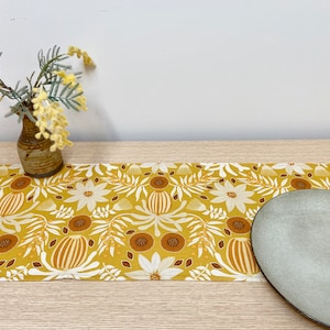Könnte beinhalten: Ein gelber Tischläufer mit einem floralen Muster mit weißen Blüten, braunen Blättern und gelben und orangefarbenen geometrischen Formen. Ein hellgrauer Keramikteller liegt auf dem Tischläufer.