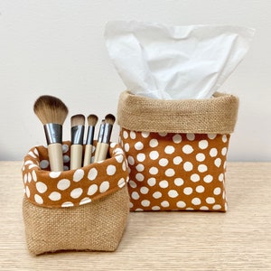 May include: Two brown and white polka dot fabric storage baskets with burlap trim. One basket holds makeup brushes and the other holds a white tissue box.