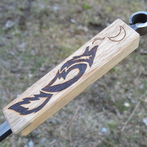 May include: A wooden bottle opener with a metal loop at the top. The wood is light brown and has a dark brown wolf design burned into it. There is a crescent moon design above the wolf.