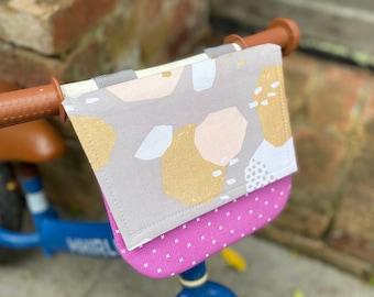 May include: A blue balance bike with a pink and grey handlebar bag with a geometric pattern. The bag has a white polka dot lining.