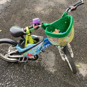 May include: A blue children's bicycle with a green basket and a matching helmet. The helmet is light blue with a dinosaur design. A purple bell is attached to the handlebars. The bike has a black seat and tyres.