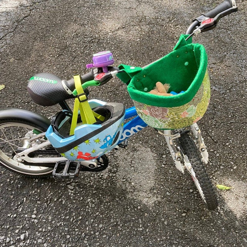 May include: A blue children's bicycle with a green basket and a matching helmet. The helmet is light blue with a dinosaur design. A purple bell is attached to the handlebars. The bike has a black seat and tyres.