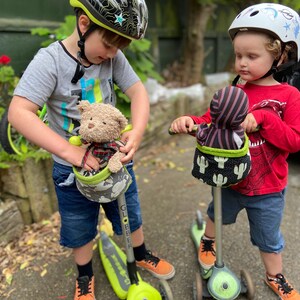 May include: Two children on scooters, each with a plush toy in a handlebar basket. One child wears a black and grey helmet with a star design, the other a white helmet with a unicorn design. The scooters are lime green and grey.