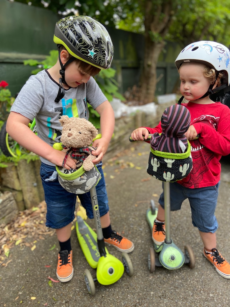 May include: Two children on scooters, each with a plush toy in a handlebar basket. One child wears a black and grey helmet with a star design, the other a white helmet with a unicorn design. The scooters are lime green and grey.