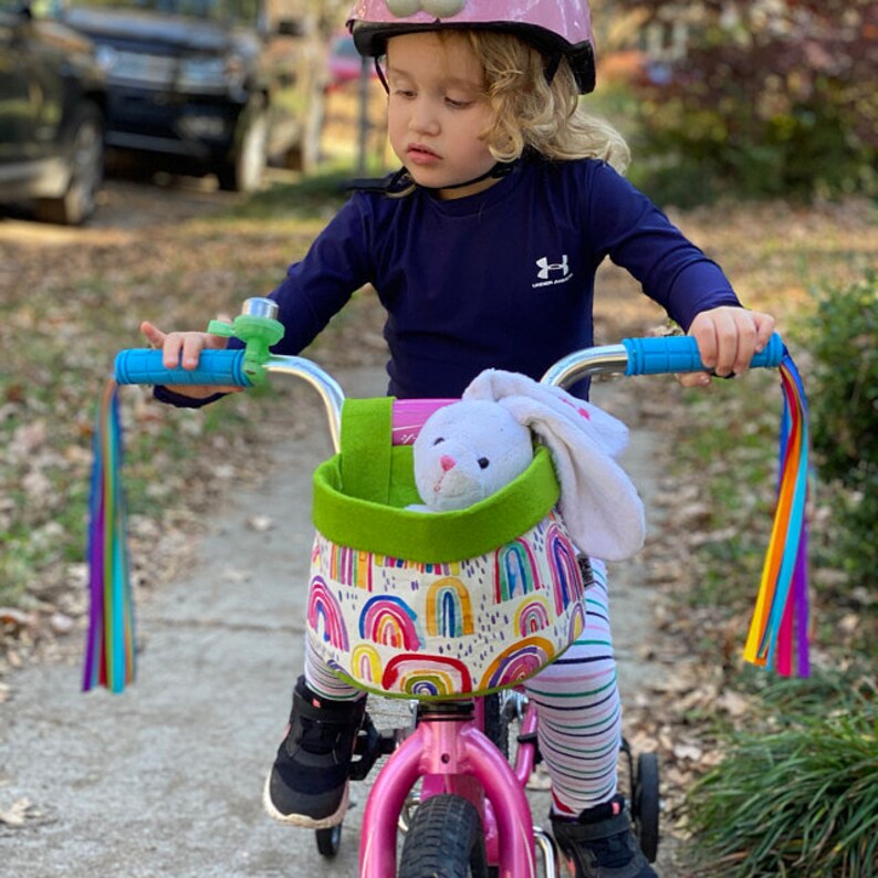 May include: A pink child's bicycle with a rainbow-patterned basket and a stuffed bunny. The basket has a green felt trim. The handlebars are blue with rainbow streamers. The child wears a pink helmet and a navy blue long-sleeved top.