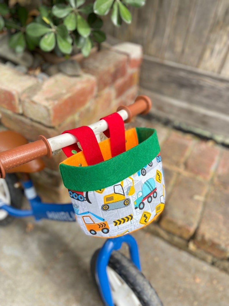 May include: A bicycle basket featuring a construction vehicle design. The basket has a green felt trim and red straps for attaching to the handlebars. The basket is attached to a blue bicycle.