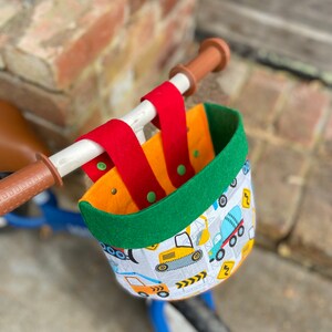 May include: A bicycle basket featuring a construction vehicle design. The basket has a green rim, red straps, and an orange interior. It is attached to the handlebars of a blue bicycle.