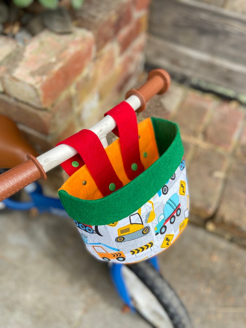May include: A bicycle basket featuring a construction vehicle design. The basket has a green rim, red straps, and an orange interior. It is attached to the handlebars of a blue bicycle.