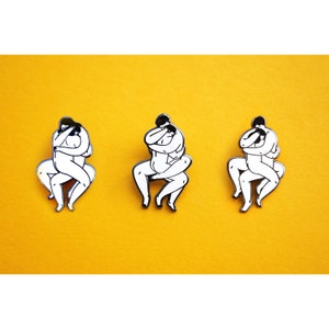 May include: Three enamel pins featuring a black and white illustration of two people embracing. The pins are on a yellow background.