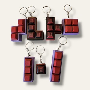 May include: Five Tetris-shaped keychains in different colors. The keychains are made of plastic and have a matte finish. The keychains are in the shape of the Tetris blocks, with each block having a different color. The keychains are attached to silver key rings.