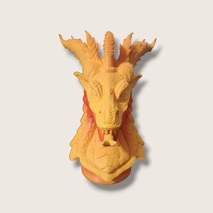 May include: A yellow dragon head sculpture with a textured surface and a red and orange gradient on the neck. The dragon has a closed mouth with a small tongue sticking out.