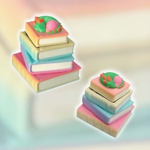 May include: Two stacks of miniature books, each featuring four volumes with pastel-colored covers in shades of pink, blue, yellow, and green. The top book in each stack is adorned with a small, sculpted green and pink decorative element.