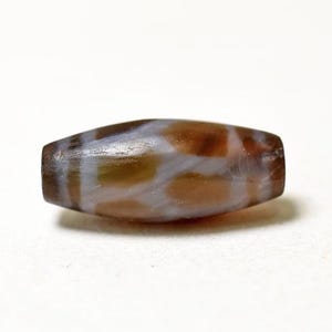 May include: A cylindrical bead with a brown and white patterned design. The bead has rounded ends and a polished surface. The pattern consists of irregular brown shapes against a white background. The bead is likely made of stone or a similar material.