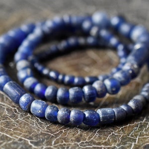 Könnte beinhalten: Eine Kette aus tiefblauen Glasperlen, wahrscheinlich aus recyceltem Glas, mit matter Oberfläche. Die Perlen sind zu einer langen, durchgehenden Kette zusammengefügt, was einen schlichten, aber eleganten Look ergibt.