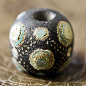 May include: A black glass bead with a decorative pattern of white and blue circles. The bead has a hole in the center for stringing.