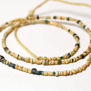 Puede incluir: Un collar compuesto por pequeñas cuentas de forma irregular en tonos beige, verde y negro. Las cuentas están ensartadas en un cordón fino y claro, creando una apariencia delicada y antigua. El collar se muestra sobre un fondo blanco.