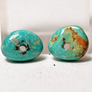 May include: Two turquoise polished stone beads with a hole drilled through the centre. The beads have a rounded, organic shape and display natural colour variations, including brown and orange veining.
