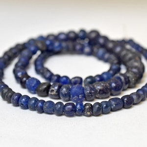 以下が含まれることがあります： 濃い青色のガラスビーズのネックレス。一部のビーズは薄い青色をしています。ビーズは円形に繋がっていて、ネックレスになっています。