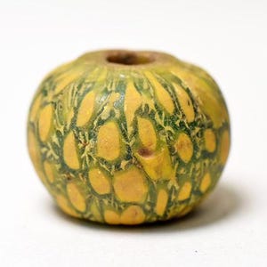 May include: A spherical bead featuring a mottled pattern of yellow and green colors. The surface has a textured appearance with irregular yellow shapes separated by a network of dark green lines. A central hole is visible at the top of the bead.