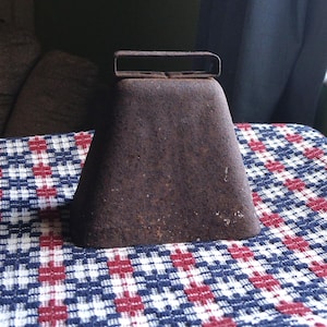 May include: A rustic, weathered metal cowbell with a rectangular handle. The bell is dark brown and sits on a patterned fabric with a red, white, and blue geometric design. The bell is a vintage item.