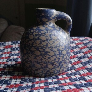 May include: A ceramic jug with a rounded body and a single side handle. The surface features a mottled pattern of dark blue speckles over a light tan base color. The jug has a short, narrow neck and a smooth, glossy finish throughout the exterior.