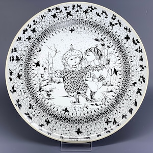 Puede incluir: Plato de cerámica blanco y negro con una escena de invierno con dos niños patinando sobre hielo. El plato tiene un borde decorativo de flores y hojas estilizadas.