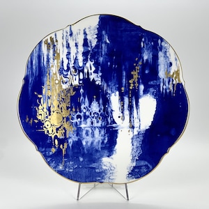 Puede incluir: Un plato de cerámica azul y blanco con detalles dorados. El plato tiene un borde festoneado y presenta un diseño abstracto.