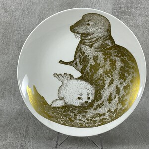 May include: A white ceramic plate with a gold design of two seals. The larger seal is on top of the smaller seal. Both seals are facing the right side of the plate.