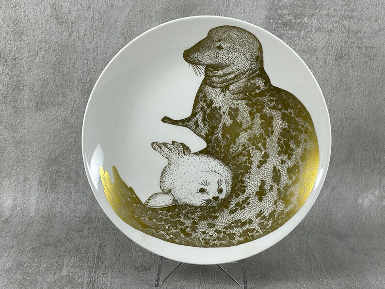 May include: A white ceramic plate with a gold design of two seals. The larger seal is on top of the smaller seal. Both seals are facing the right side of the plate.